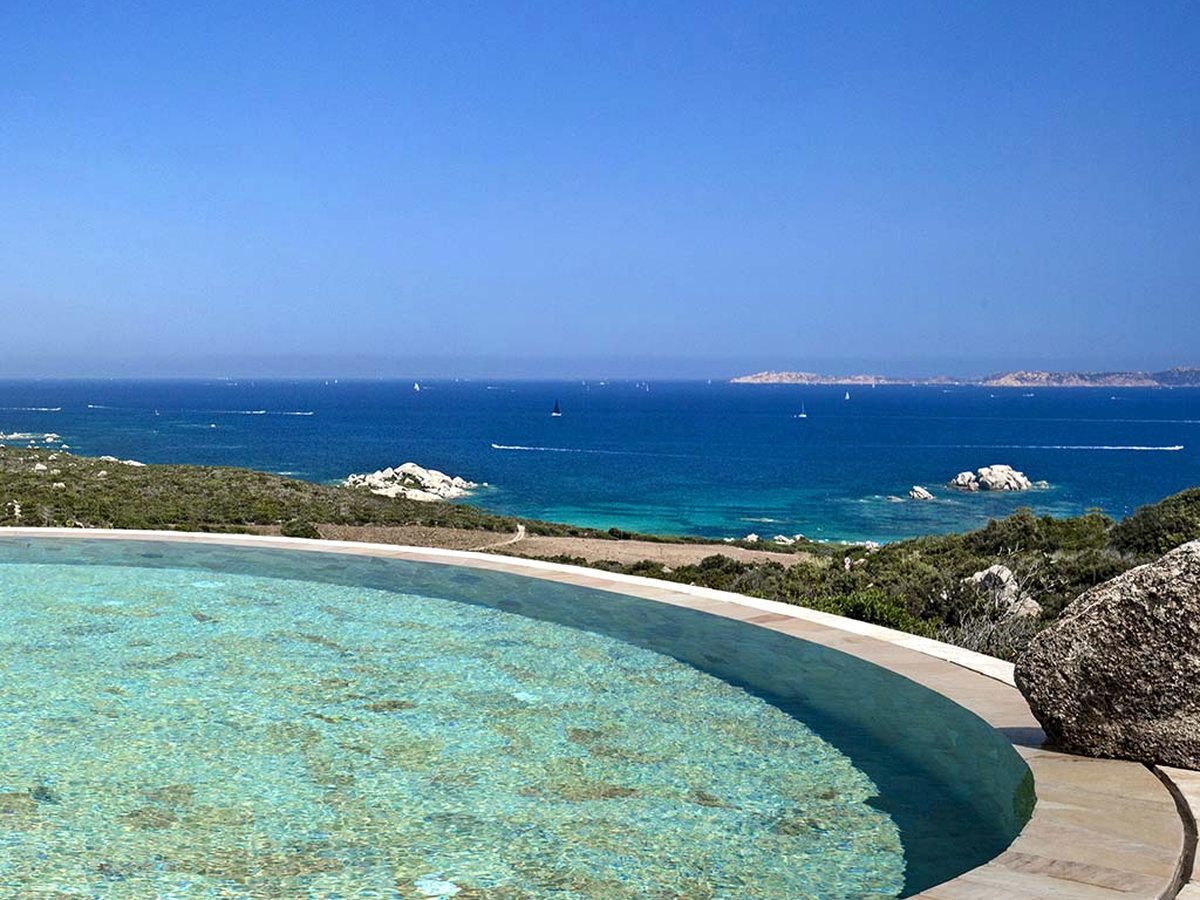 A curved pool with a stone surround in the foreground, looking over low scrub to a deep blue sea with sailing boats and rocky islands on the horizon.