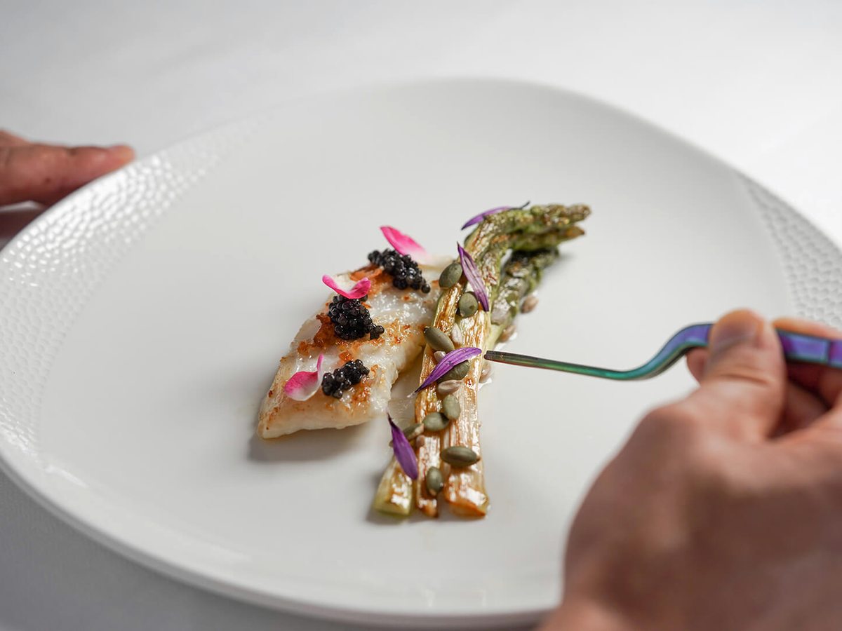 A white plate holding a fillet of fish with asparagus, caviar and edible flowers, with a hand lifting a forkful.