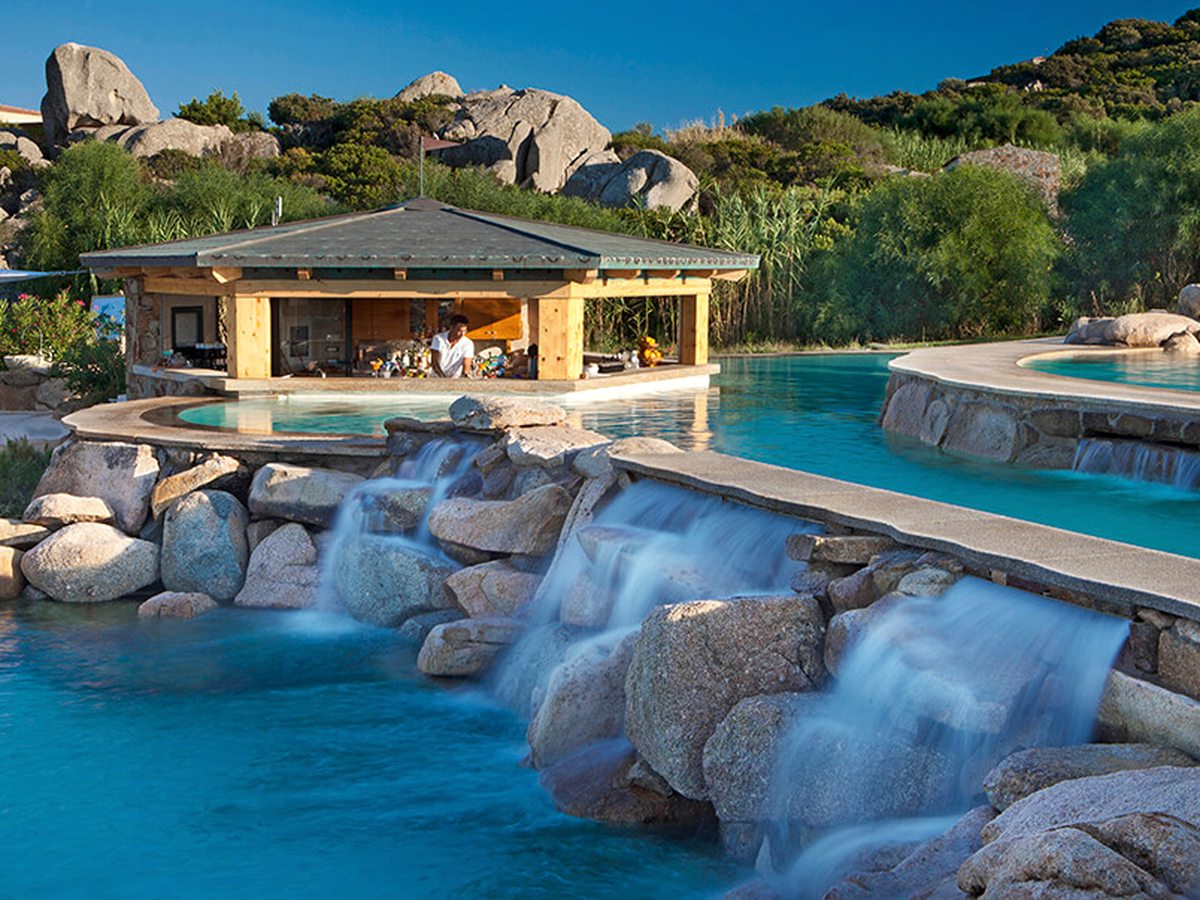 A multi-level turquoise pool with water spilling over granite boulders into the level below, and a timber-framed bar hut with a barman standing behind the counter at the water's edge.