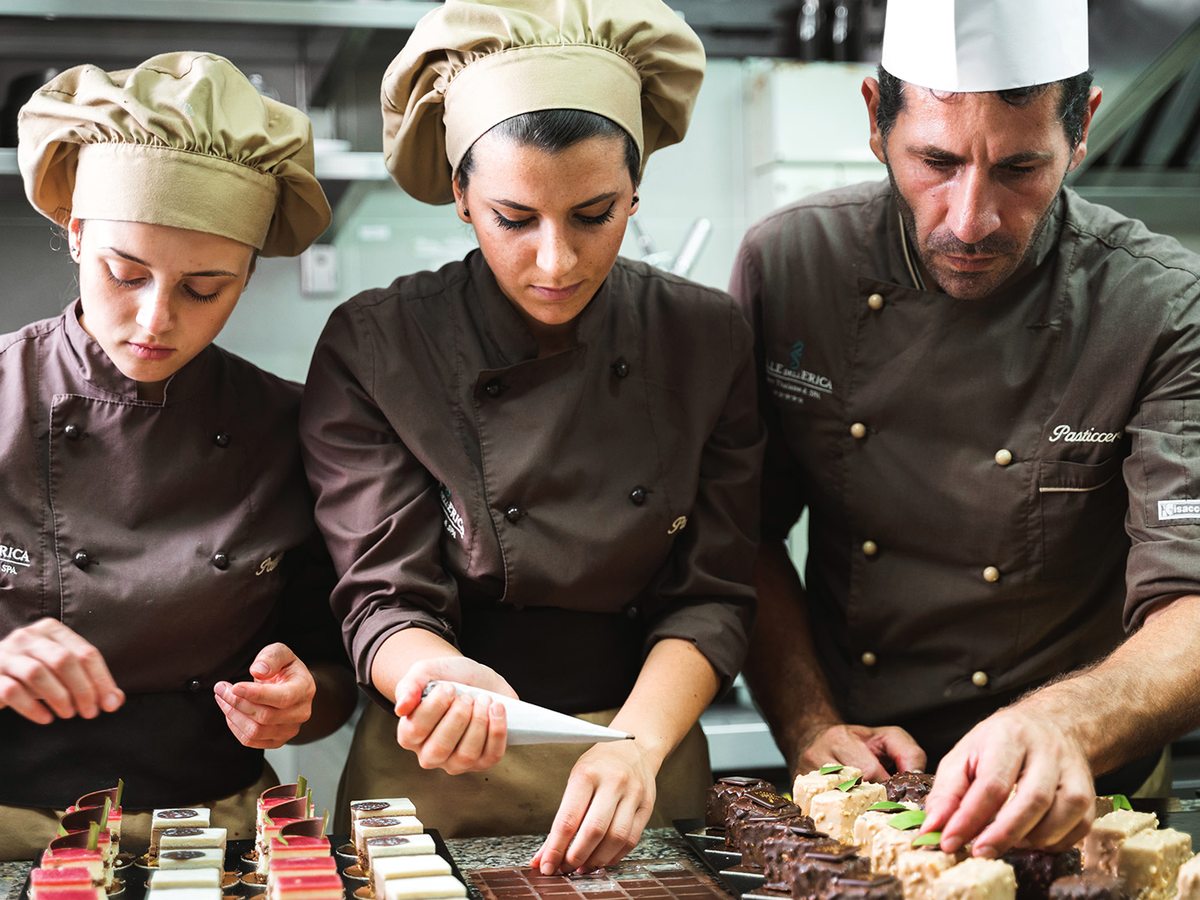 Three chefs in brown jackets and tall white hats arranging rows of small desserts along a counter.