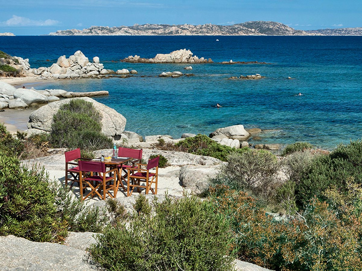 A wooden table with four red director's chairs standing on a flat granite slab above a small sandy cove, with clear turquoise water and islands on the horizon.