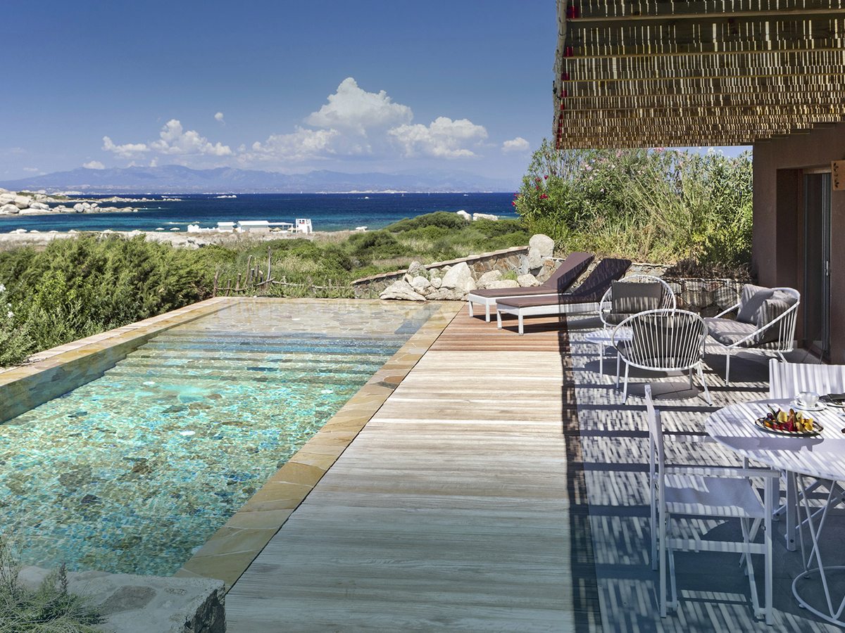 A long narrow pool beside a timber deck with sun loungers, chairs and a table laid with fruit, screened by a reed blind, with scrub, a beach and the sea beyond.