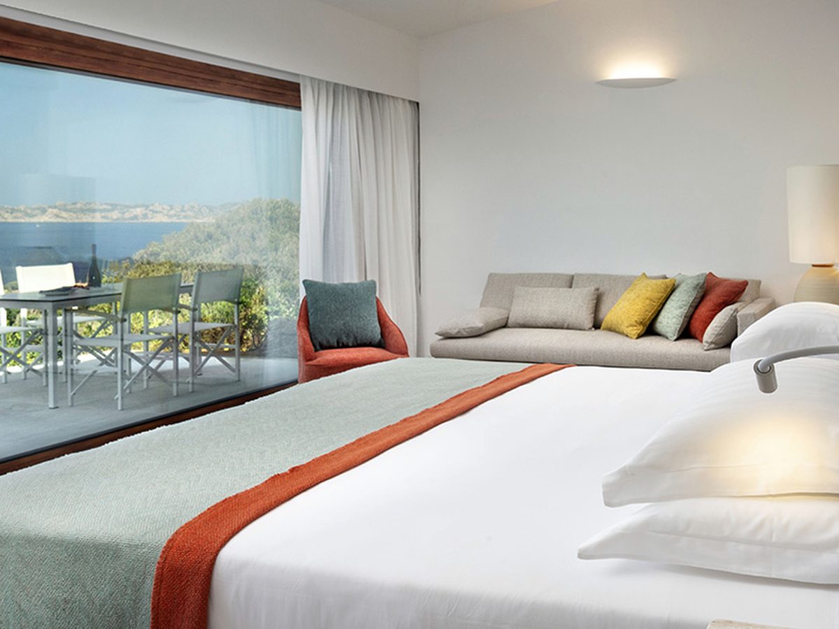 A double bed with a white cover and orange runner, a long pale sofa against the far wall and full-height glass doors onto a veranda with a table and chairs, the sea beyond.
