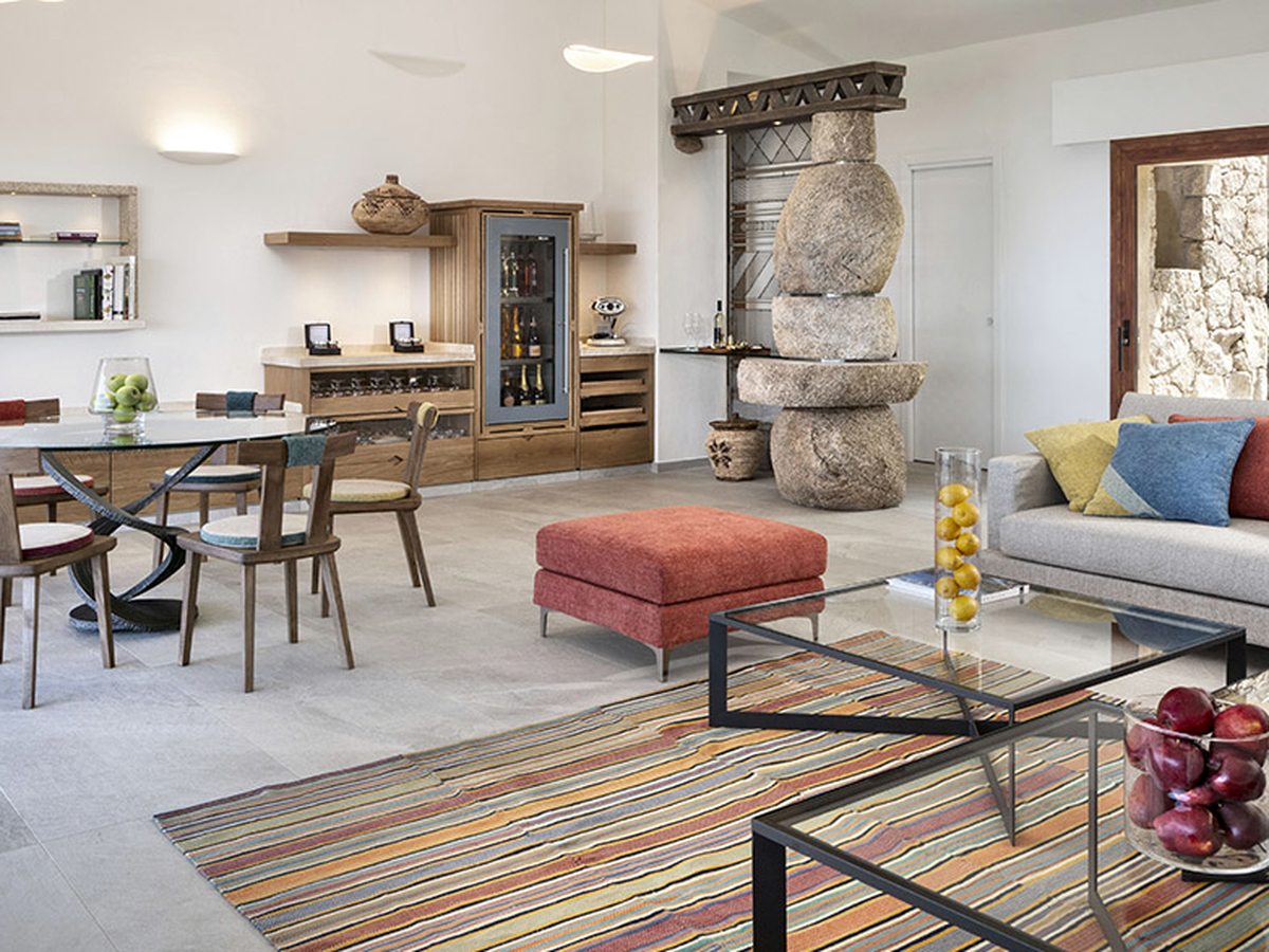 An open living and dining room with a round dining table and wooden chairs, a pale sofa, a striped rug, open shelving and a stacked granite feature by the wall.