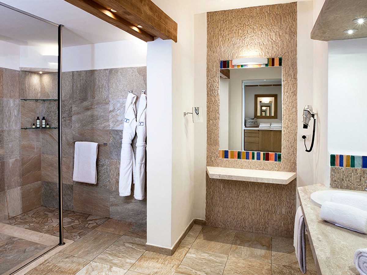 A bathroom with a glass walk-in shower, a single oval basin on a stone counter, a mosaic-framed mirror above a stone shelf, folded towels and white robes on hooks.