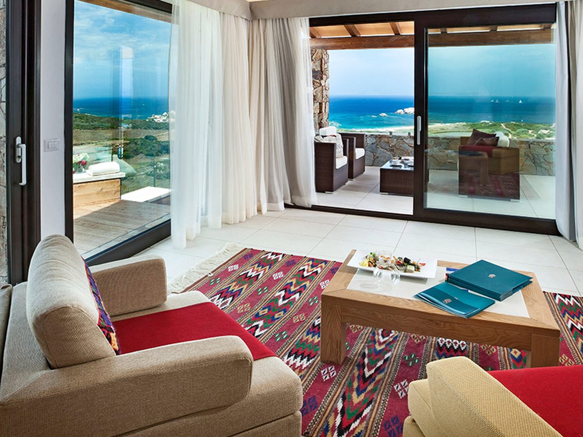 A living room with pale sofas, red throws and a patterned rug, with sliding glass doors onto a covered terrace with wicker armchairs and a low table, and the sea beyond.