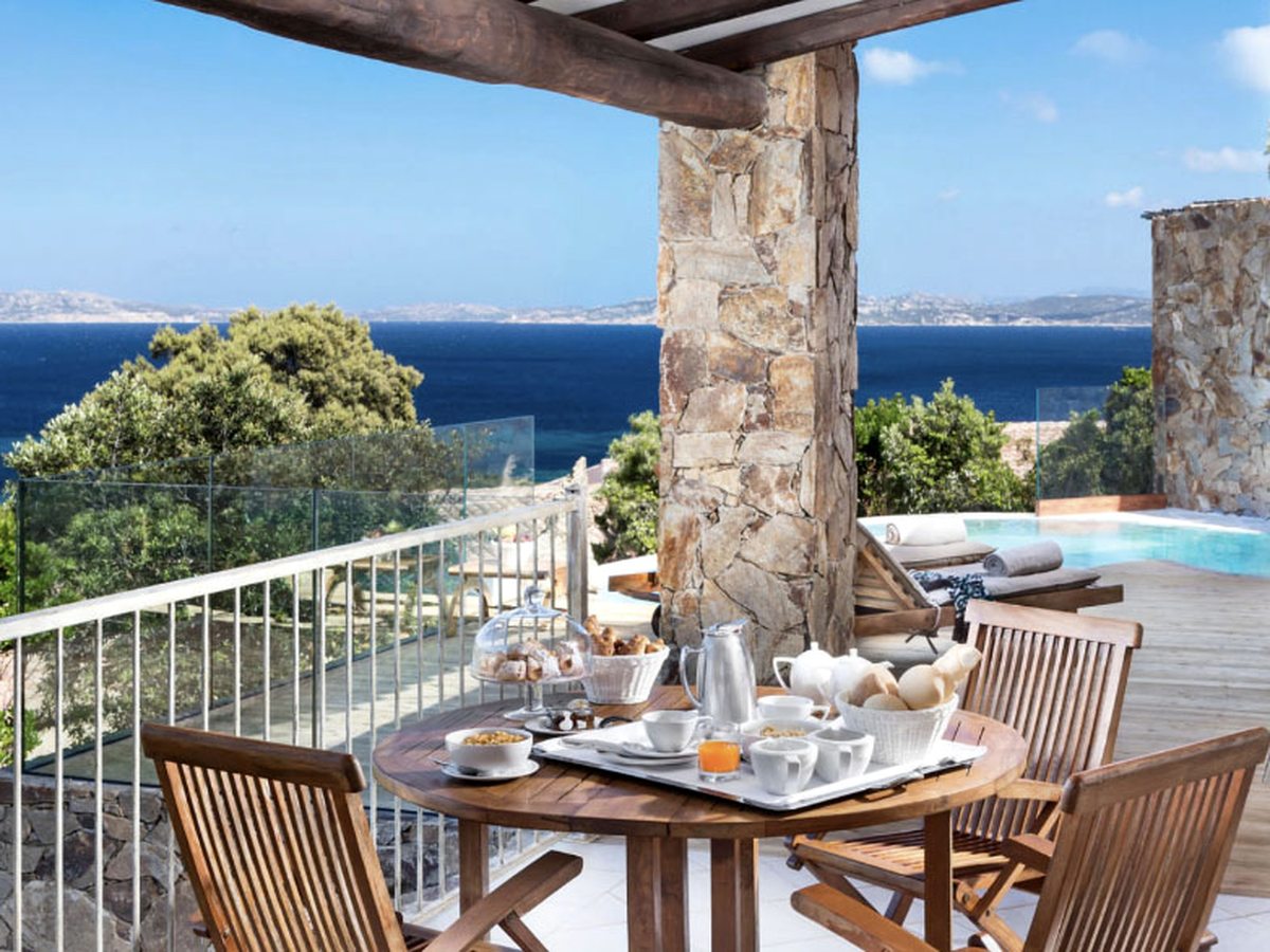 A covered veranda with a round wooden table laid for breakfast, folding wooden chairs, a stone pillar and a glass balustrade, with a small pool to one side and the sea in the distance.