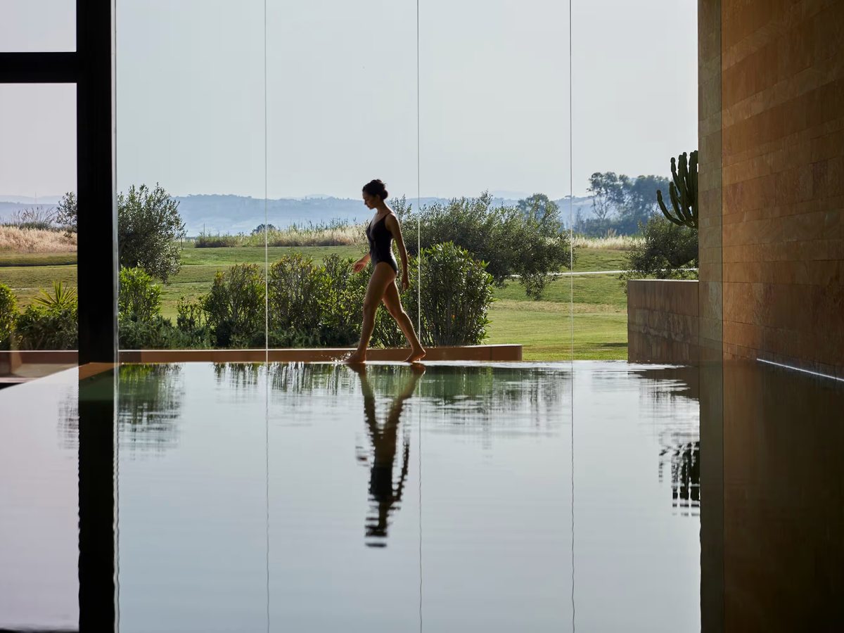 Long indoor pool with a mirror-still surface beside a full-height glass wall looking out to olive trees, the golf course and hills.