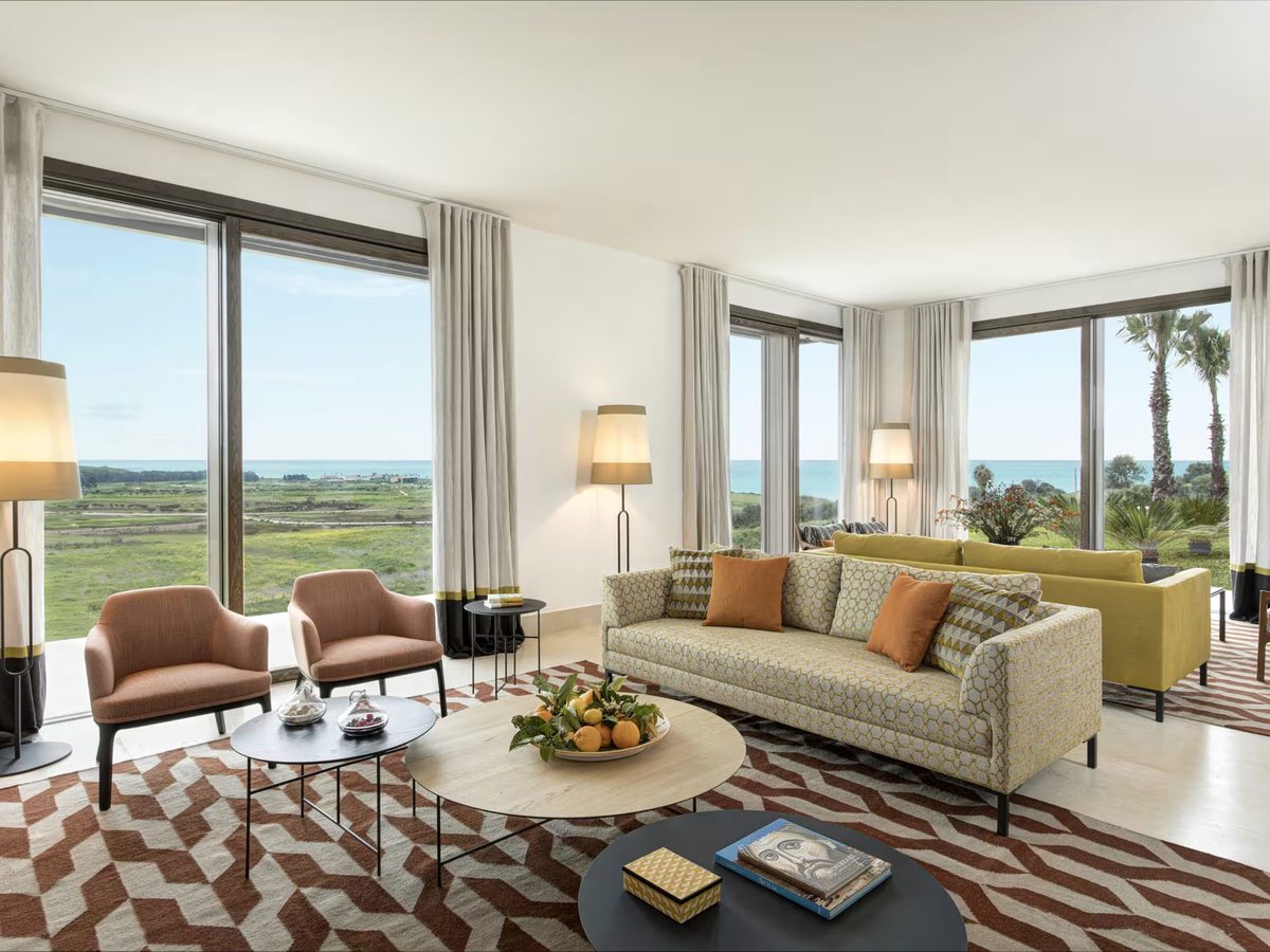 Bright villa living room with a patterned sofa, pink armchairs, a green sofa and floor lamps on a geometric rug, with floor-to-ceiling corner windows over the grounds and the sea.