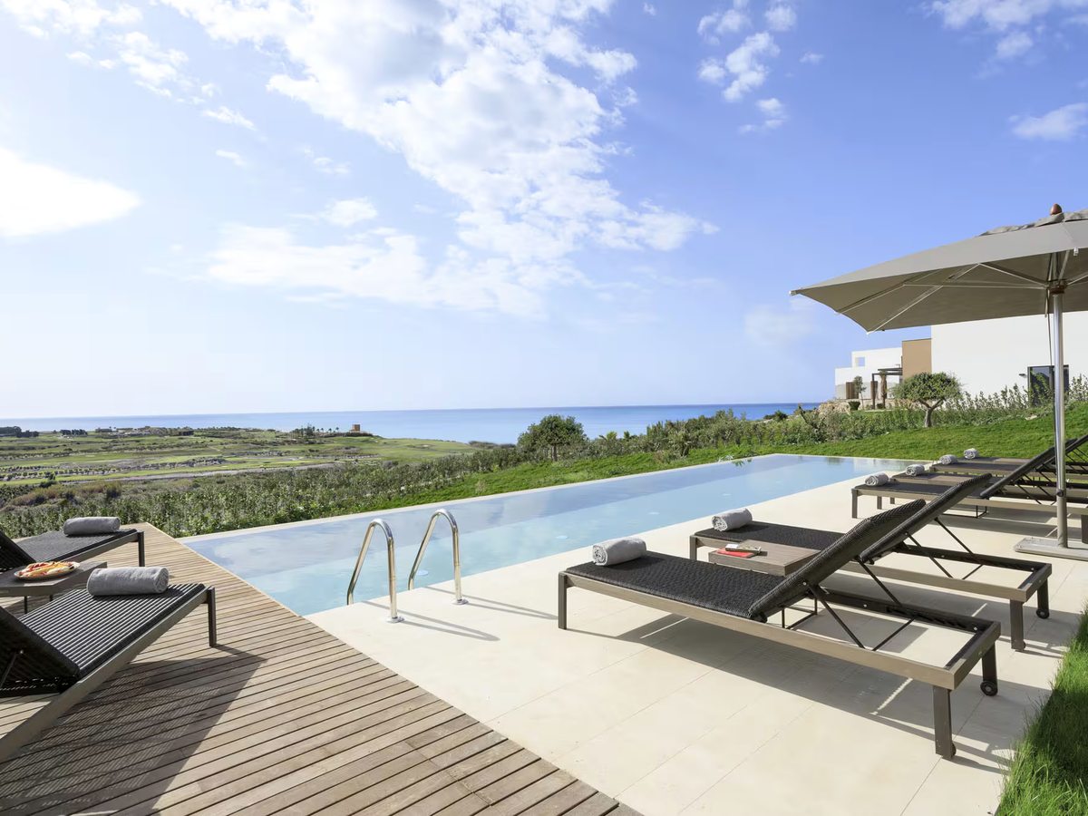 Narrow private infinity pool with steel steps, dark sun loungers and a parasol on a timber and stone terrace, with the golf course and sea beyond.