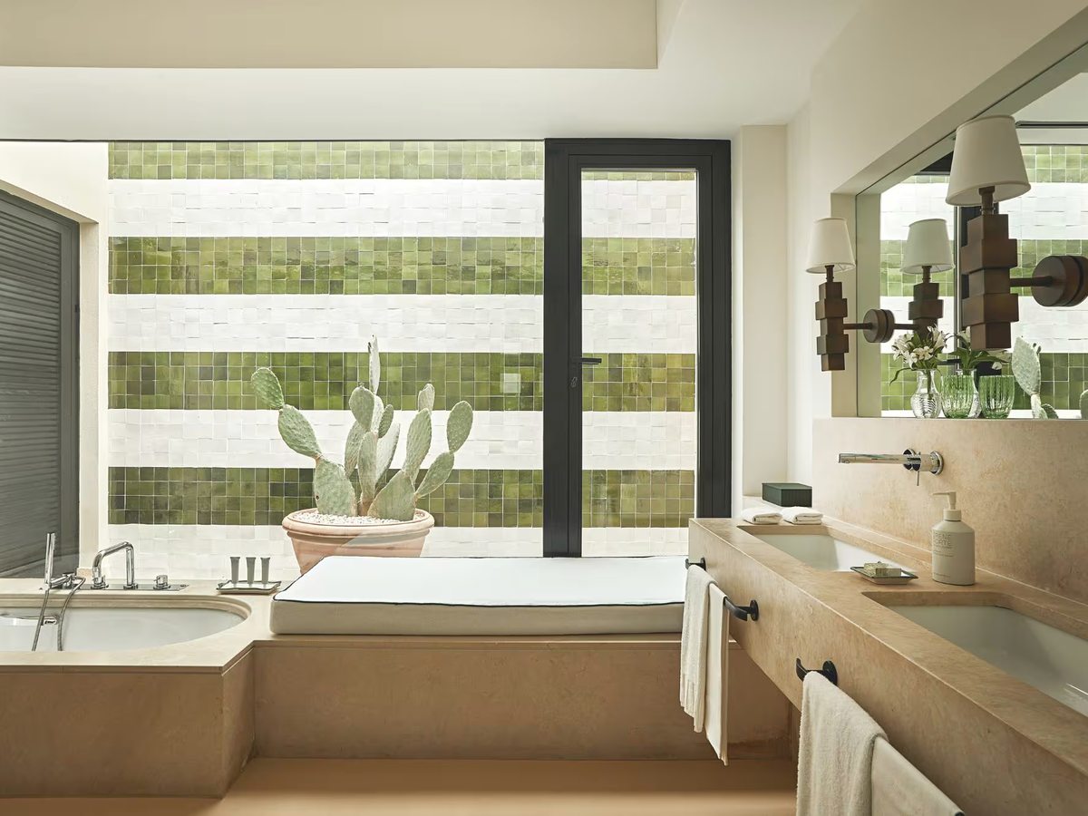 Stone bathroom with a built-in bath, a long twin-basin vanity, wall mirrors and green and white tiled walls with a potted cactus.