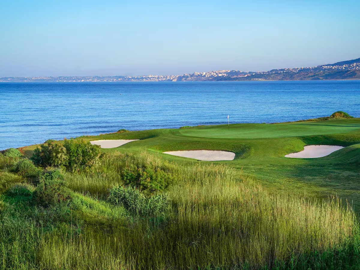 Golf green with a white flag and three pale sand bunkers set among rough dune grasses above a calm blue sea, with a distant hillside town.