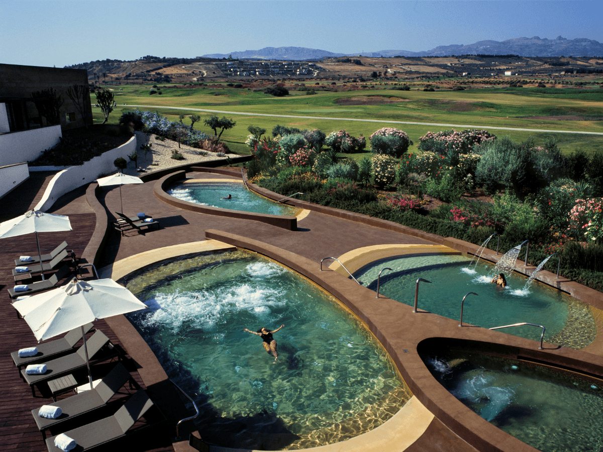 Four linked outdoor pools with bubbling water and jets, edged in terracotta decking with cream parasols and sun loungers, backed by flowering shrubs and the golf course.