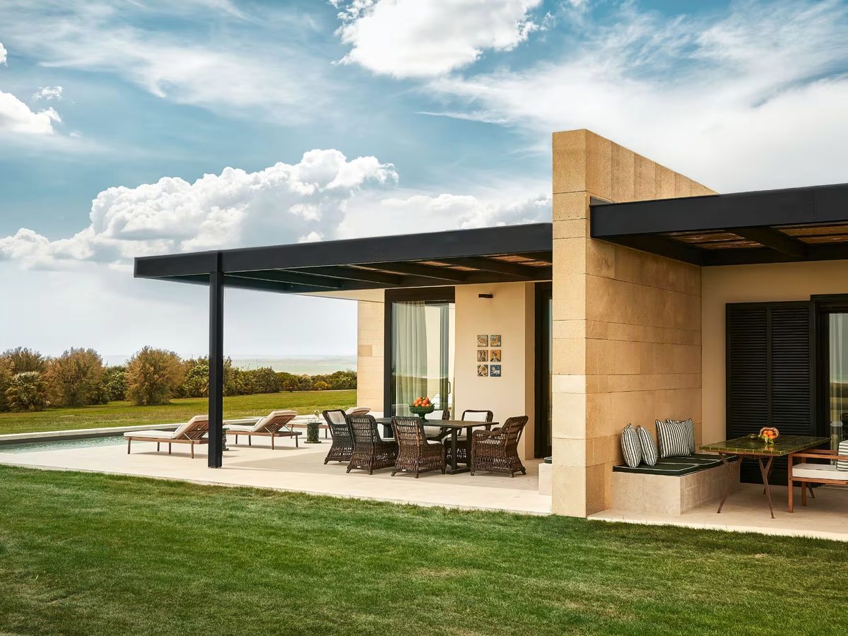 Single-storey stone-clad suite with a deep shaded terrace, wicker dining chairs and sun loungers beside a private pool on a mown lawn.