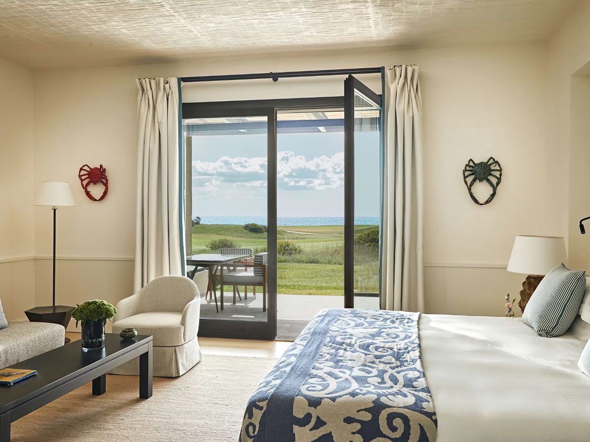 Cream bedroom with a blue and white patterned bed runner, an armchair and open glass doors onto a terrace looking over a golf fairway to the sea.