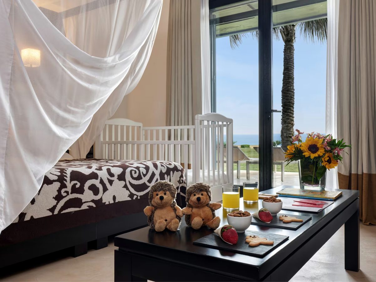White wooden cot beside a four-poster bed draped in muslin, with two hedgehog soft toys, juice and biscuits on a low table and a palm and the sea through the window.