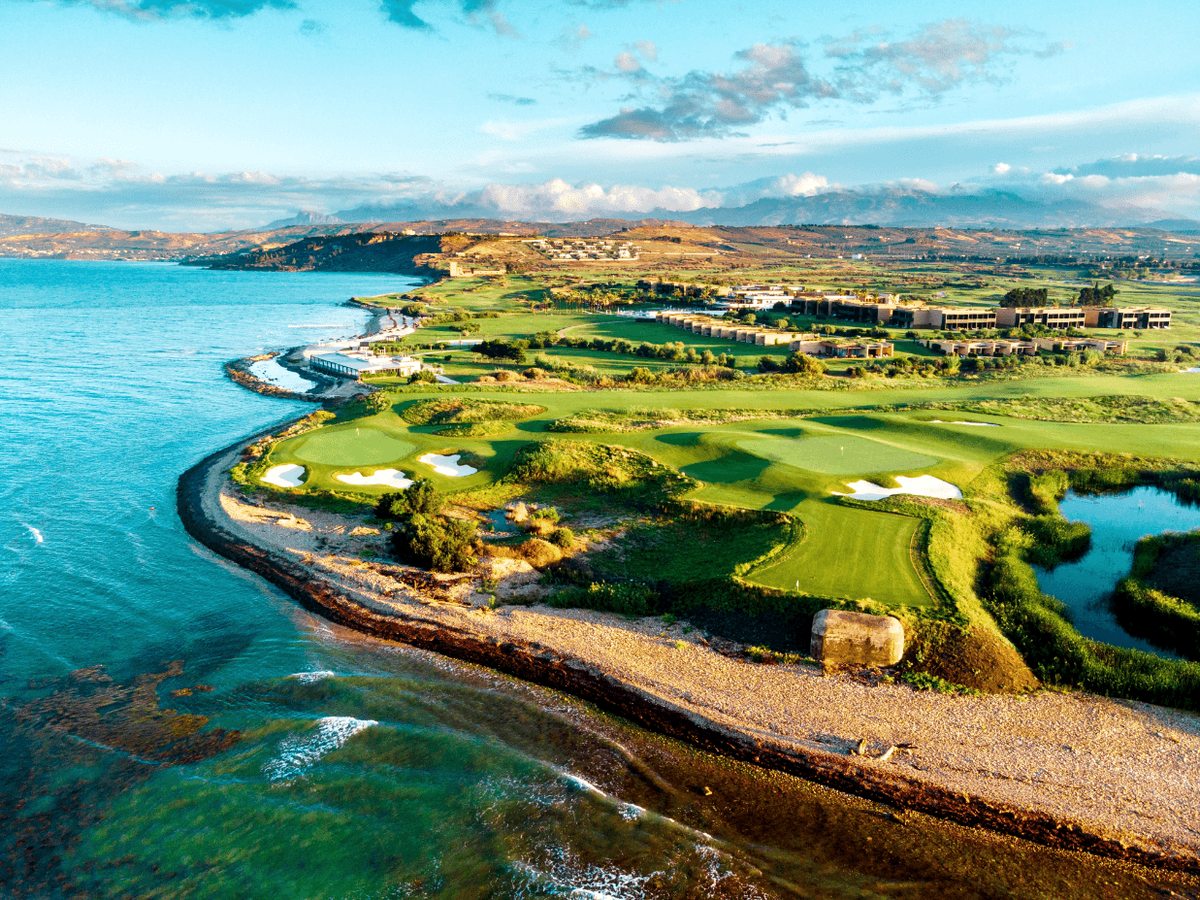 Aerial view of a green coastal golf estate, with low resort buildings, fairways and greens running along a turquoise sea and hills inland.