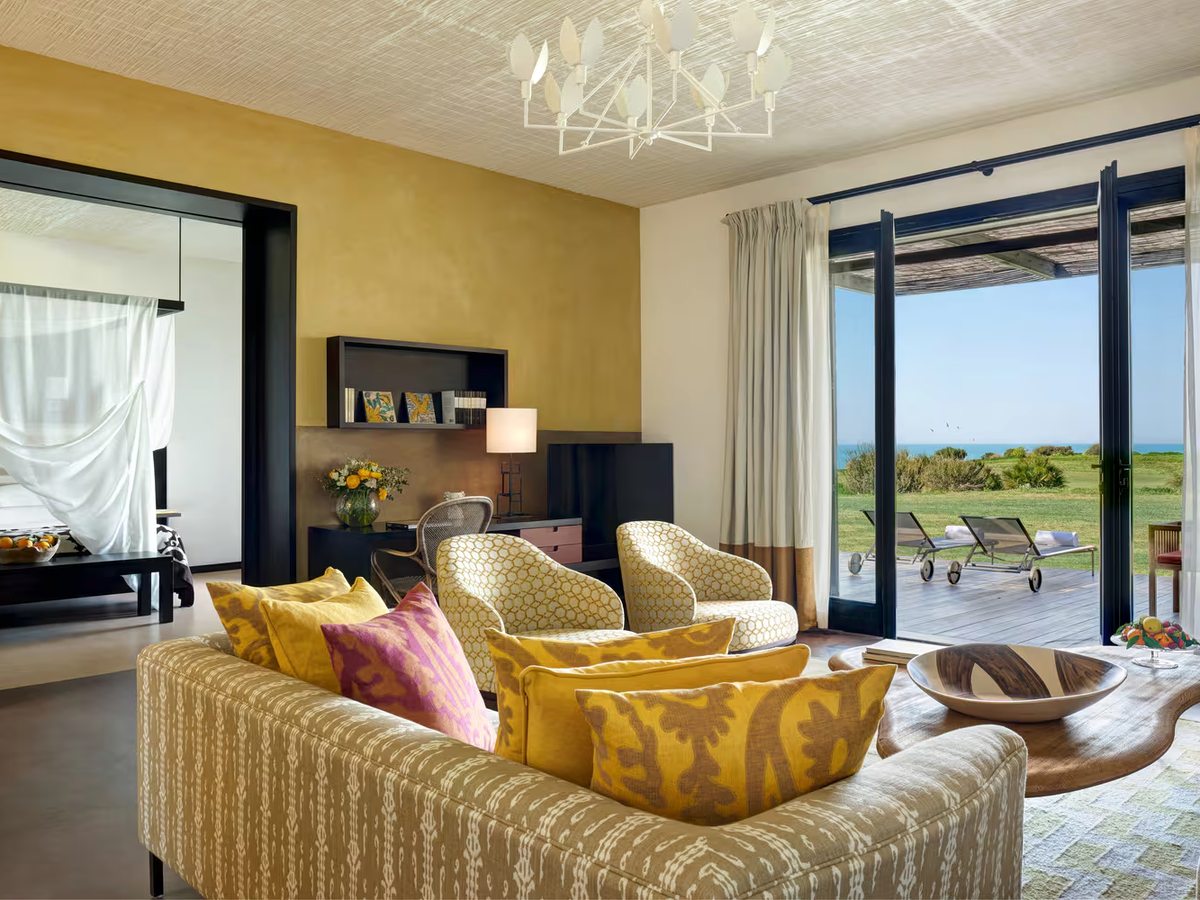 Suite living room with a patterned sofa, yellow cushions and armchairs under a white chandelier, folding glass doors onto a decked terrace and a four-poster bed visible through a doorway.