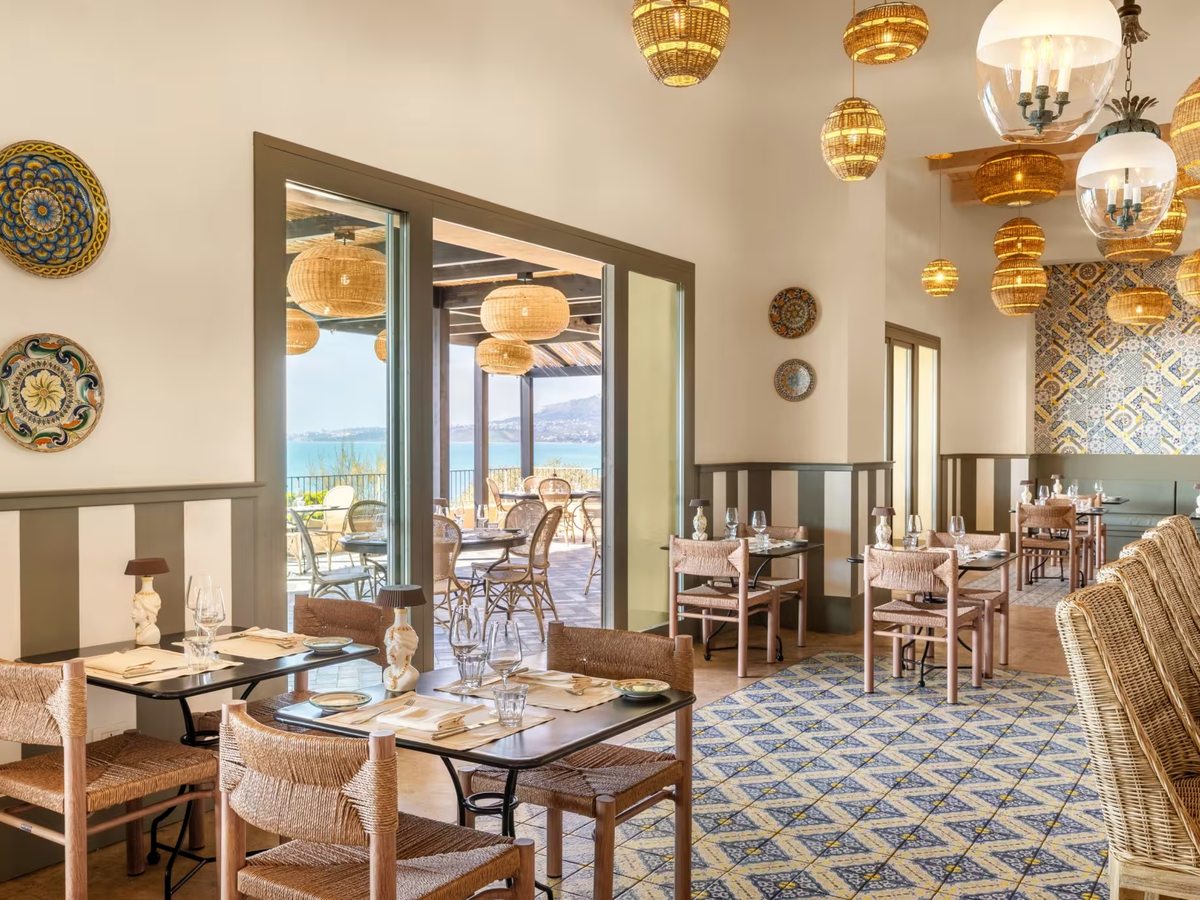 Cream dining room with patterned blue and yellow floor tiles, woven rush chairs, hand-painted ceramic plates on the walls and rattan pendant lights, opening to a sea-view terrace.