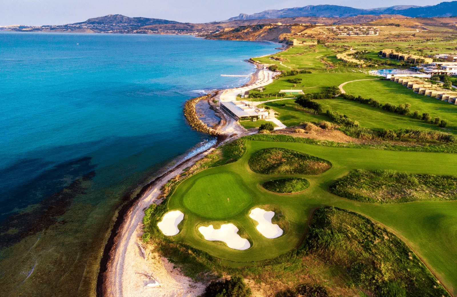 Verdura Resort on the south-west coast of Sicily, low sand-coloured buildings and golf courses running along a turquoise bay, with the resort beach and its parasols on the shore.