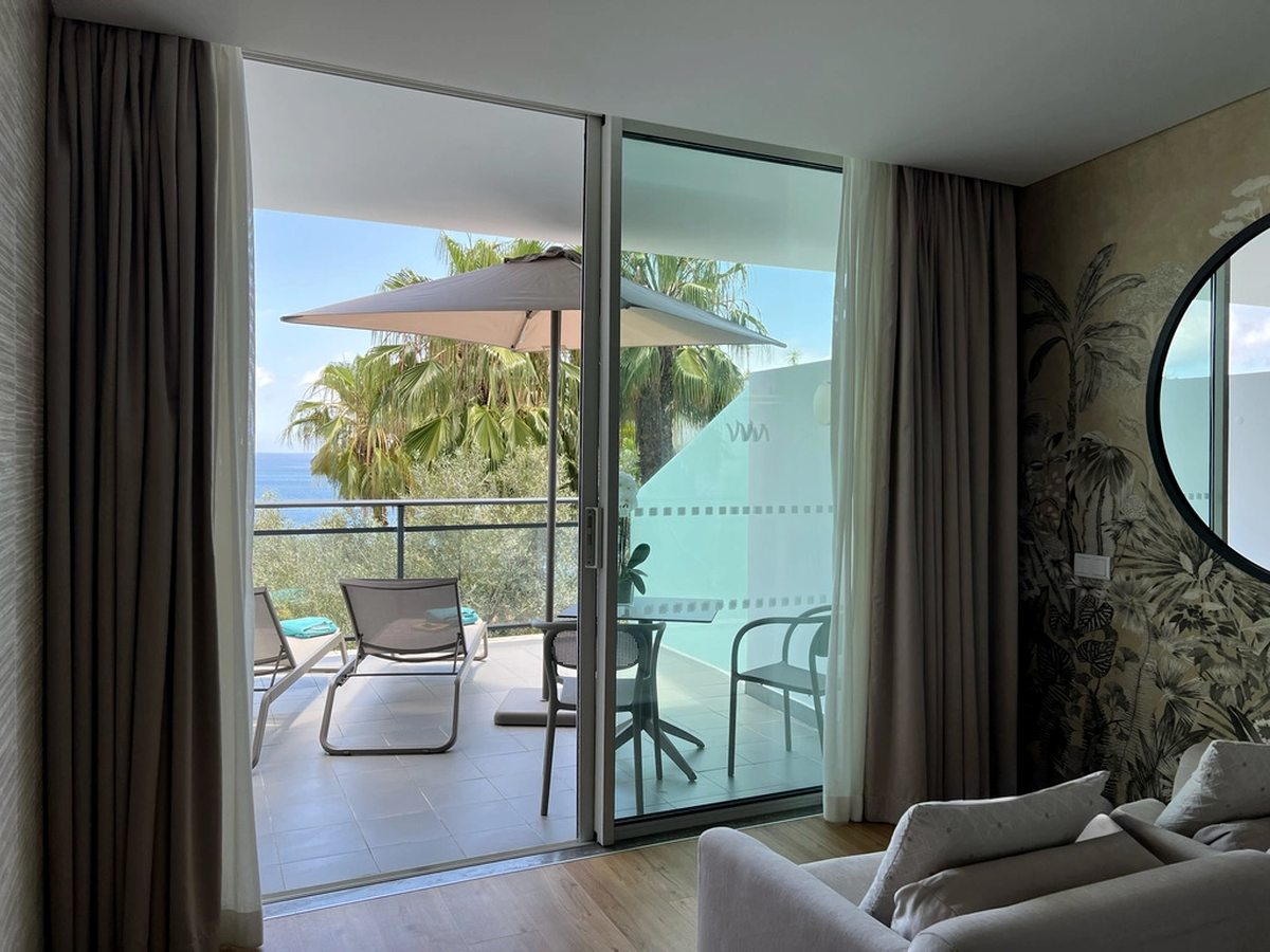 A large tiled private patio with two sun loungers, a parasol and a table and chairs behind a glass screen, with palms and the sea beyond.