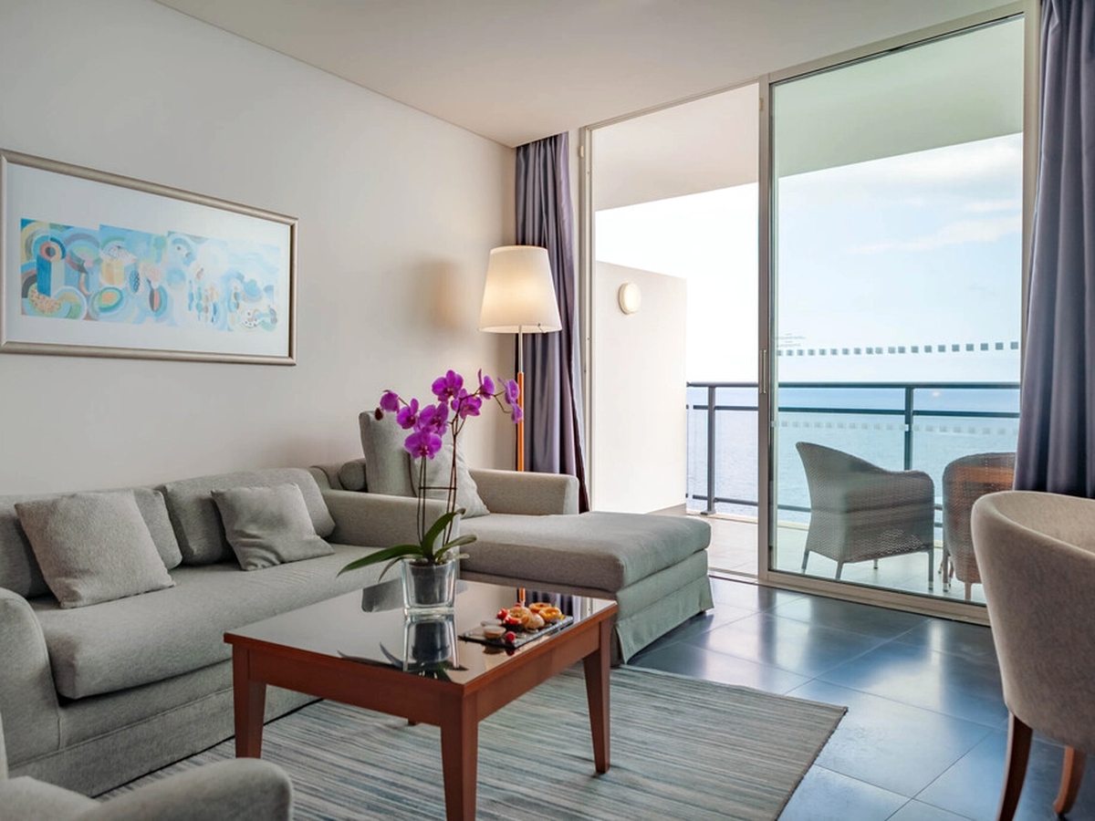 A suite living area with a grey corner sofa, a wooden coffee table and an orchid, opening through glass doors onto a balcony above the sea.
