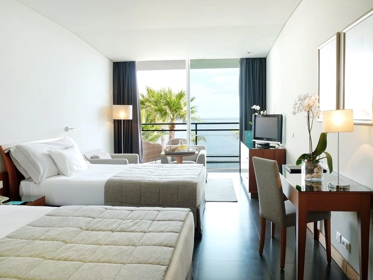 A room with two single beds side by side, a wooden desk and chair, a television and a balcony with wicker chairs and palms beyond.