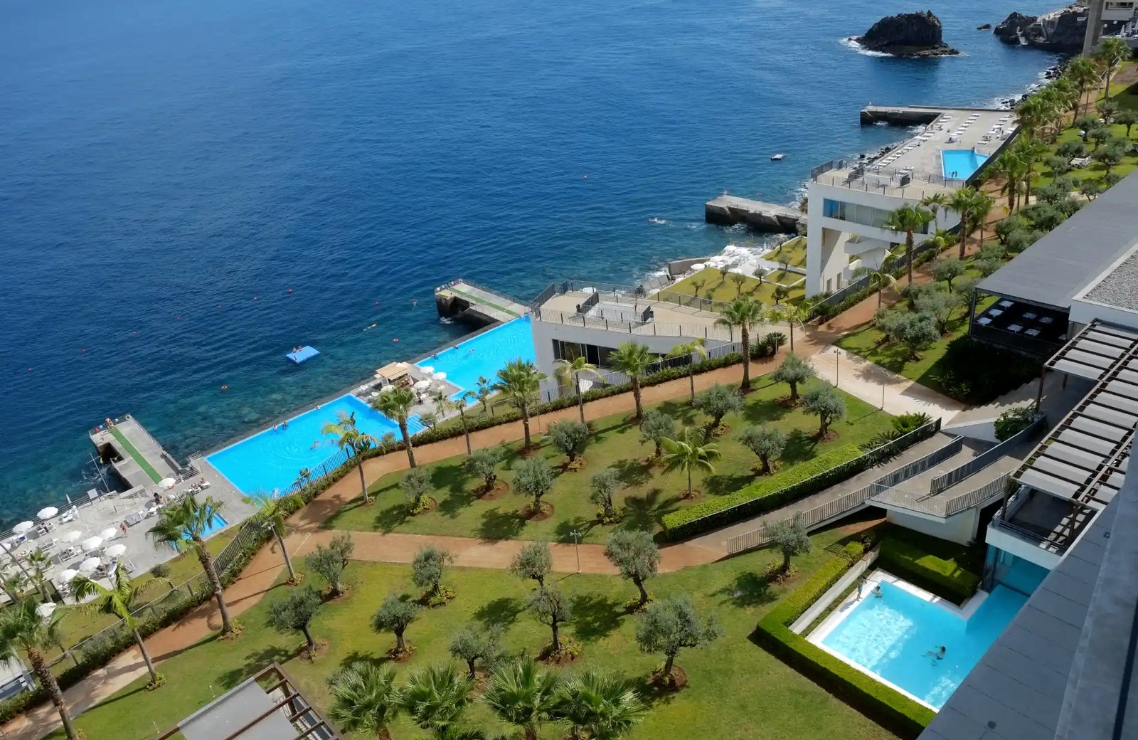 VidaMar Resort Madeira from above: blue swimming pools on terraces beside lawns of olive and palm trees, with a concrete sea platform reaching into the Atlantic below the hotel buildings.