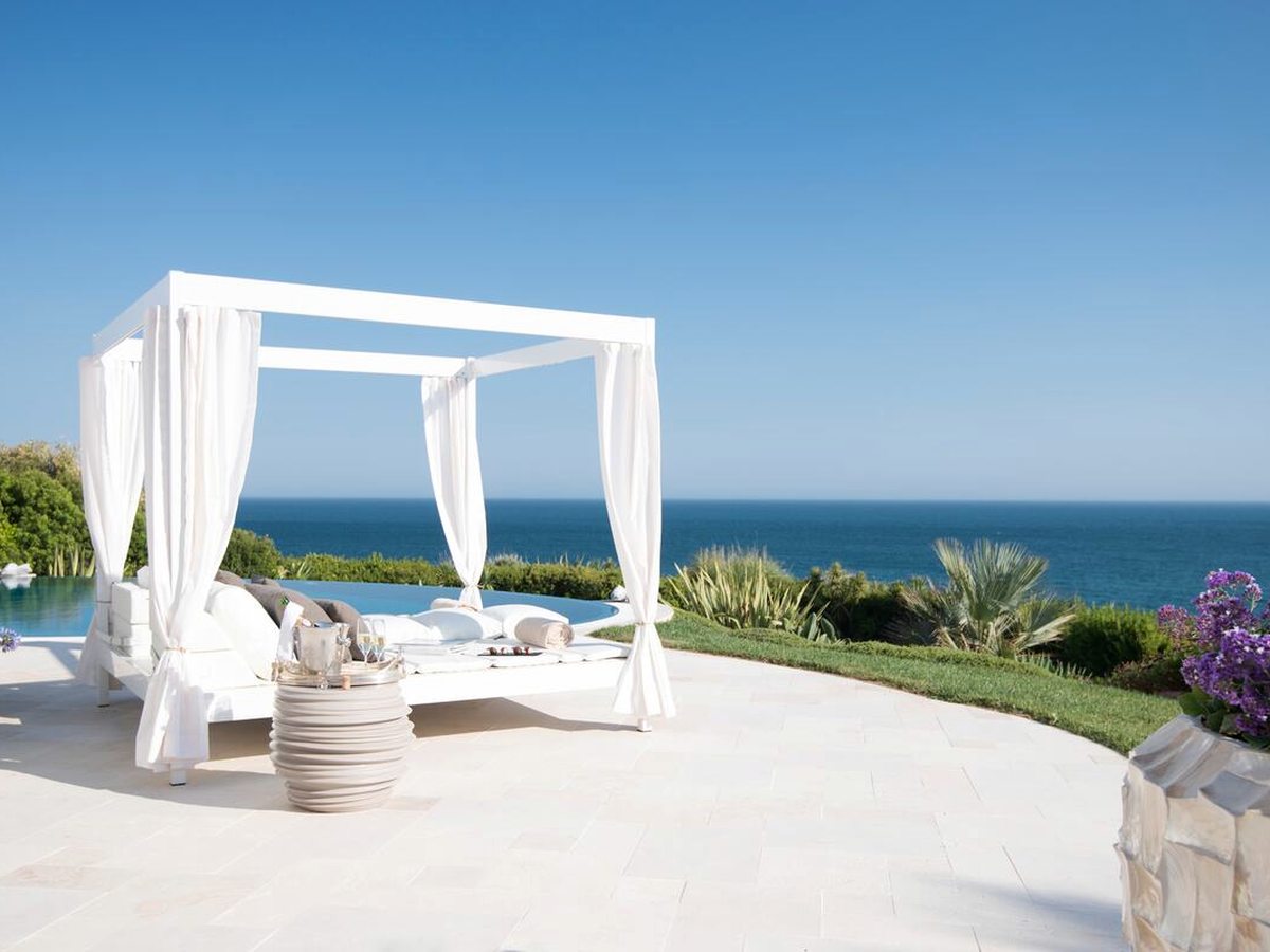 A curtained white four-poster daybed on a pale stone terrace beside an infinity pool, with flowering shrubs, palms and the Atlantic beyond.