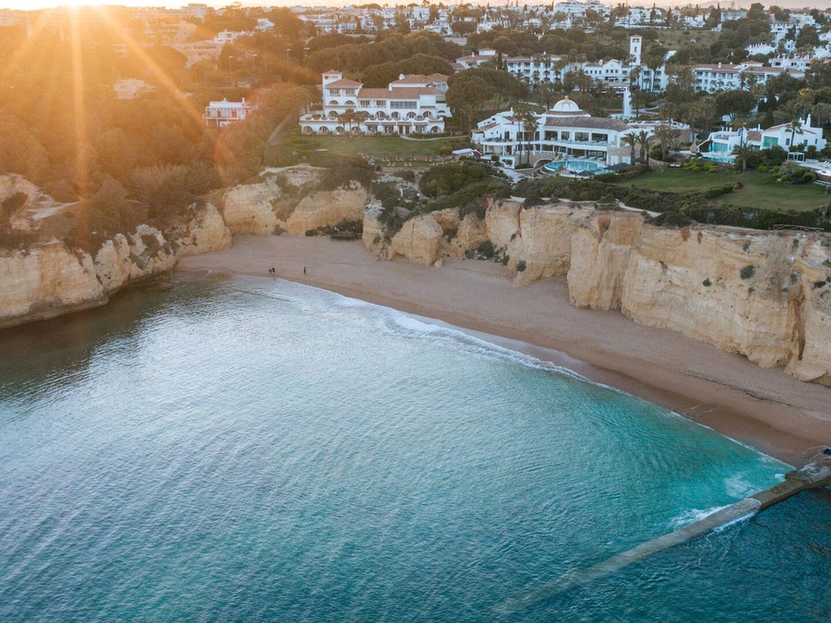 An aerial view of a curved sandy cove below ochre cliffs, with the resort's white buildings, lawns and a pool along the clifftop above it.
