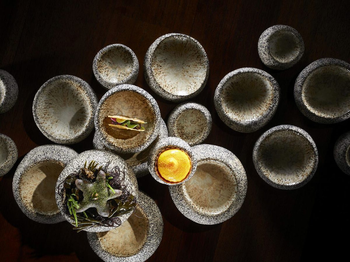 A cluster of small speckled ceramic bowls on a dark wooden table, three of them holding a garnished bite and the rest empty.