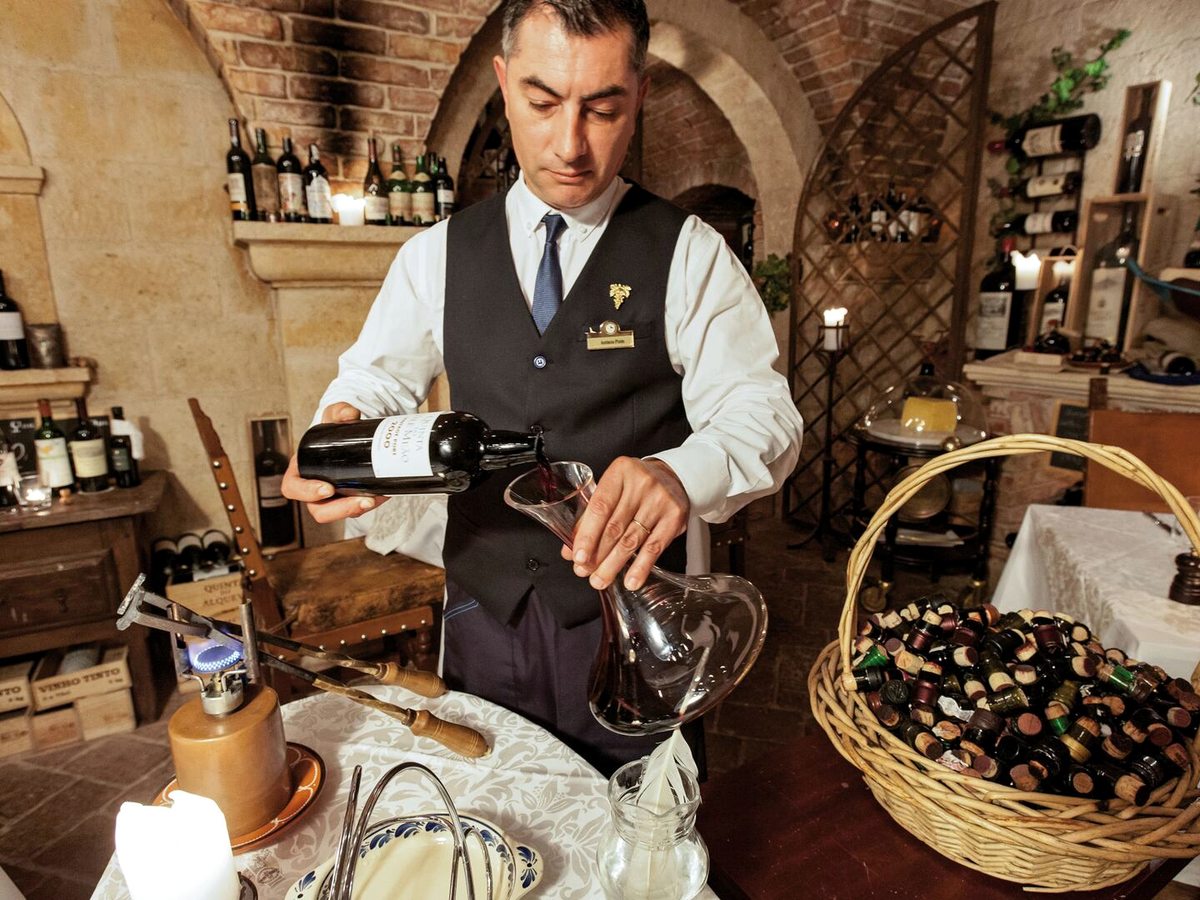 A sommelier in a waistcoat decanting red wine at a candlelit table in a brick-vaulted cellar lined with wine bottles and a basket of corks.