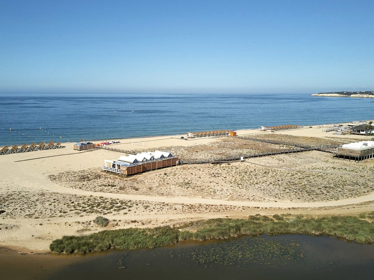 An aerial view of a long flat sandy beach with rows of thatched parasols and timber beach restaurants, backed by dunes and a lagoon.