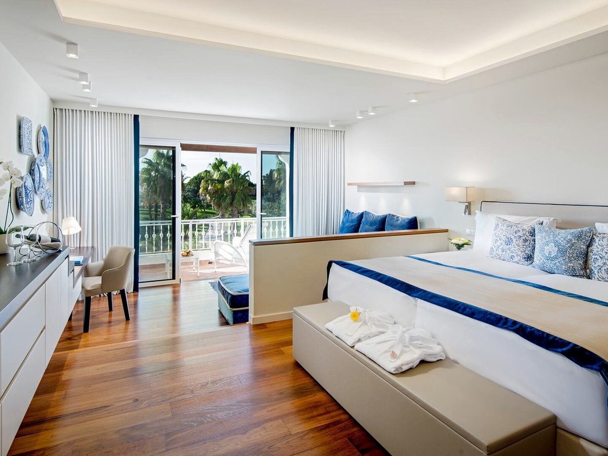 A bright room with a wide bed dressed in white and blue, a dark wood floor, a desk along one wall and balcony doors open to palms and lawn.