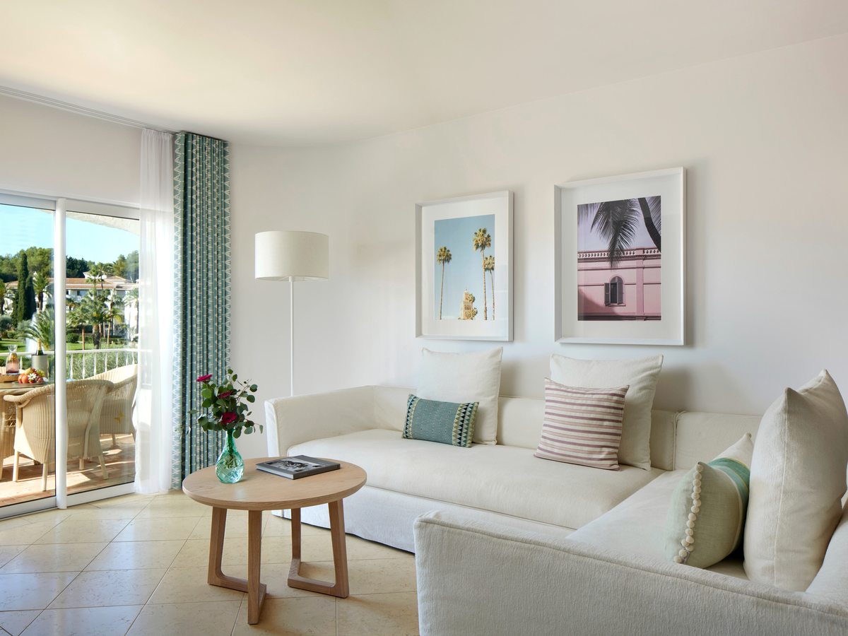 A cream lounge with two large sofas, a round wooden coffee table, framed prints and sliding doors onto a balcony above the gardens.