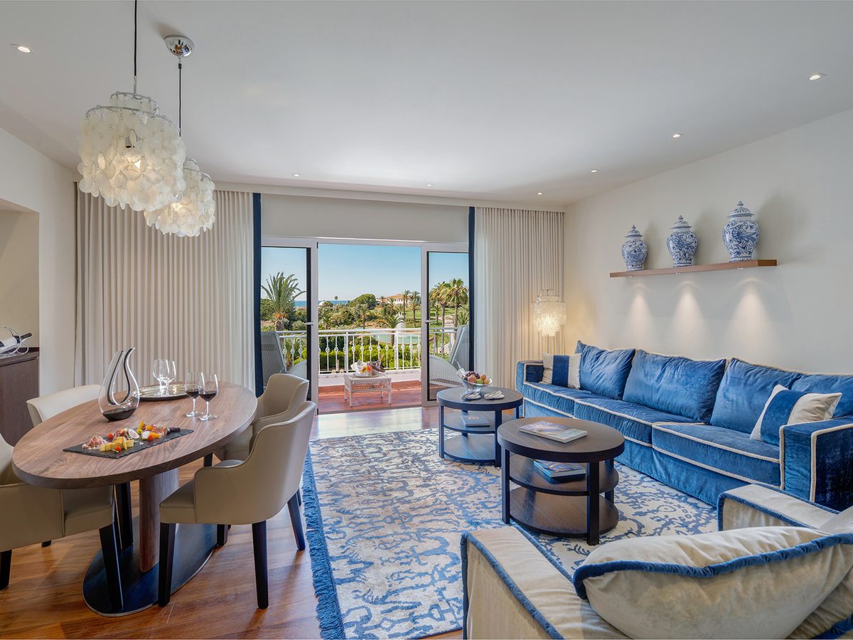 A suite living room with a long blue velvet sofa, a round dining table, a shell chandelier and balcony doors open to gardens and the sea.