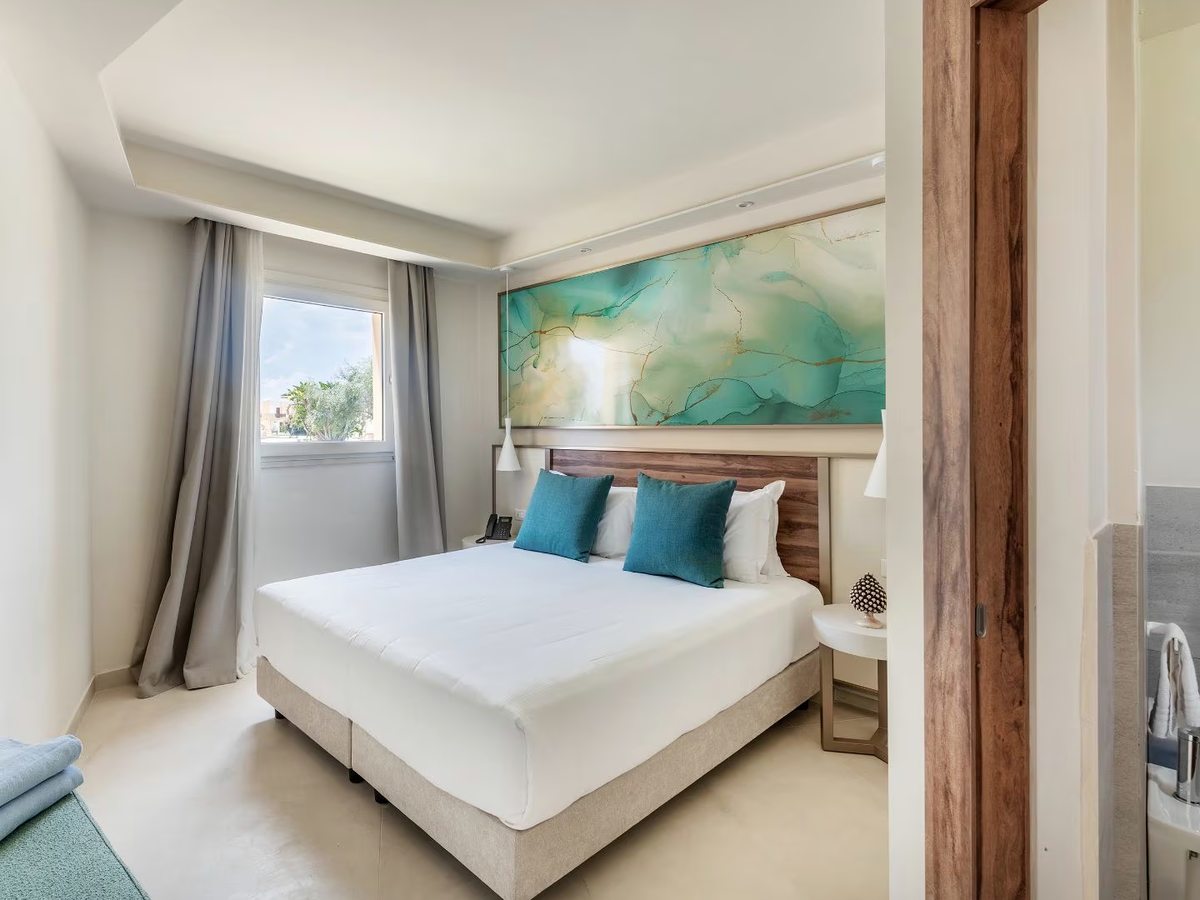 A double bedroom with a dark wood headboard, turquoise cushions and a wide turquoise abstract panel above the bed, a curtained window to the left and an open doorway through to the bathroom on the right.