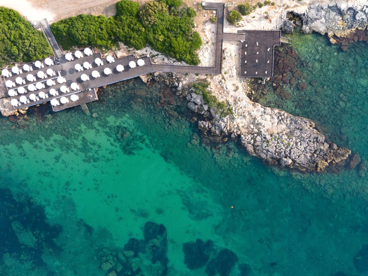 Wooden sun decks lined with rows of white parasols along a rocky shore, with a rocky spit reaching out into clear turquoise water.