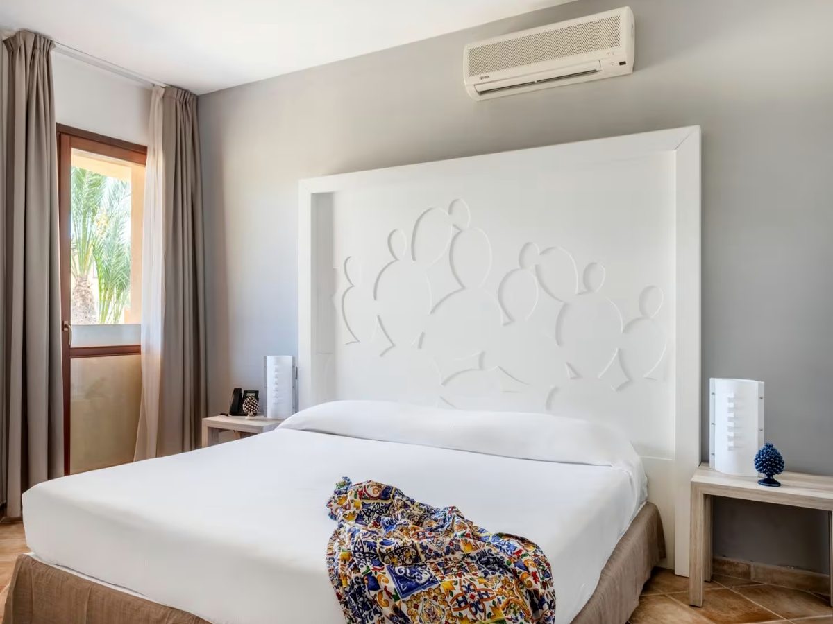A double bedroom with a tall white headboard moulded with a prickly-pear pattern, a colourful Sicilian-print throw across the bed, pale bedside tables and a glazed door onto a terrace with palms outside.