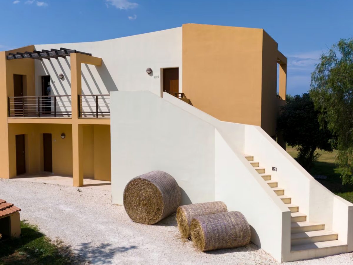 A two-storey guest villa rendered in ochre and white, with ground-floor room doors under an upper balcony and an external staircase, standing on pale gravel among trees under a blue sky.