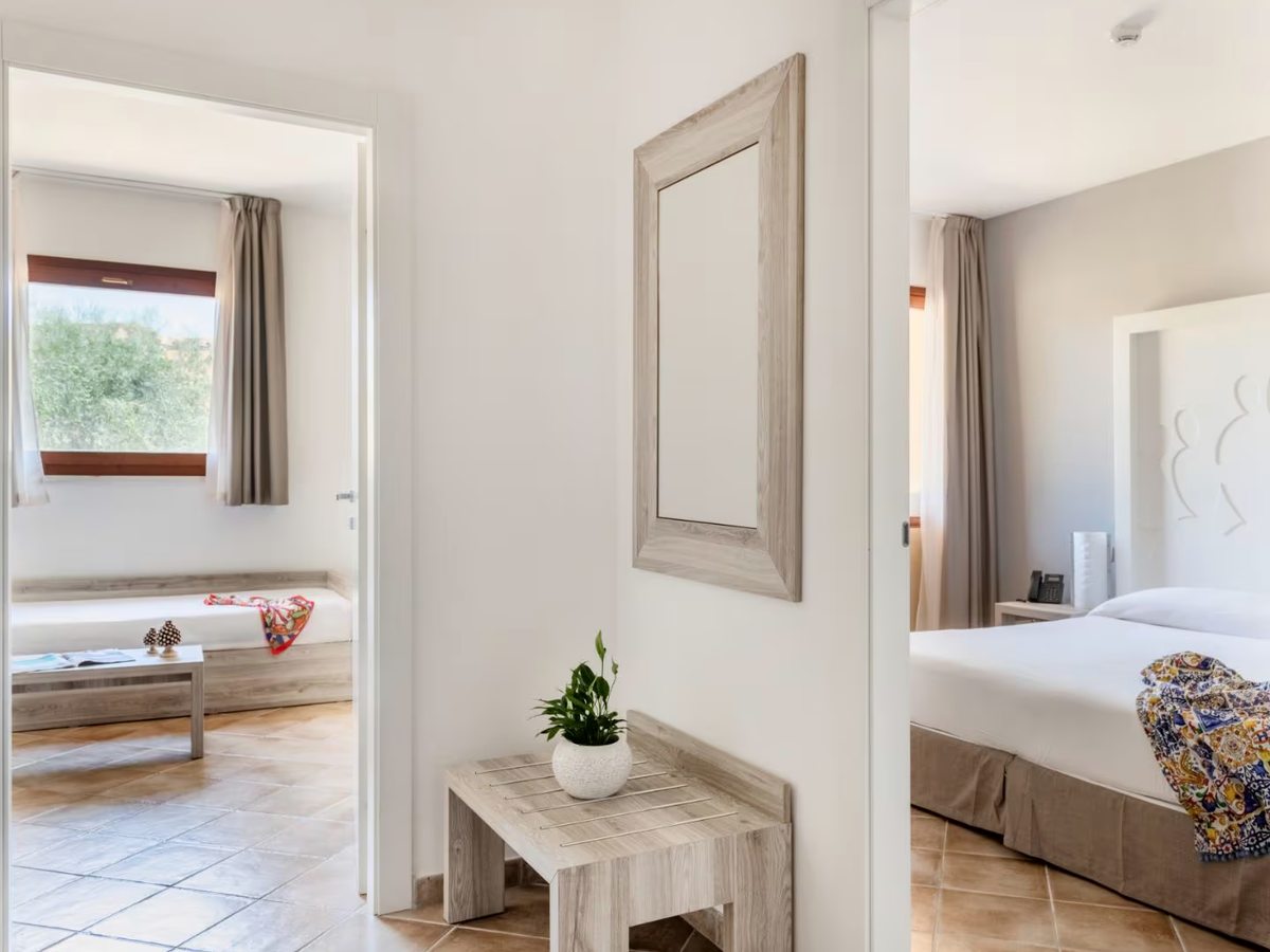An internal hallway with a mirror and a small bench, an open doorway on the left into a living room with a single bed and a doorway on the right into a double bedroom.