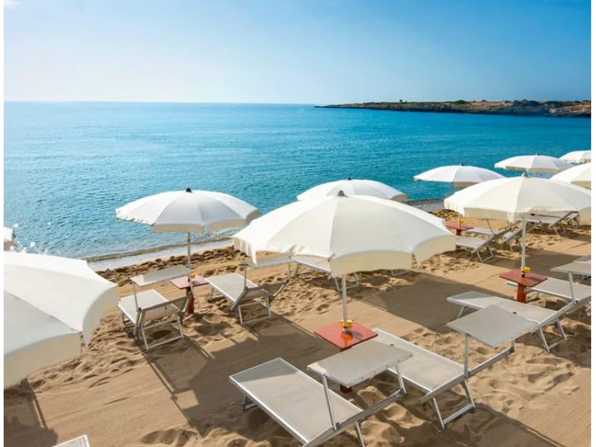 Rows of white parasols and sun loungers set out on golden sand above a calm turquoise sea, with a low rocky headland along the far side of the bay.