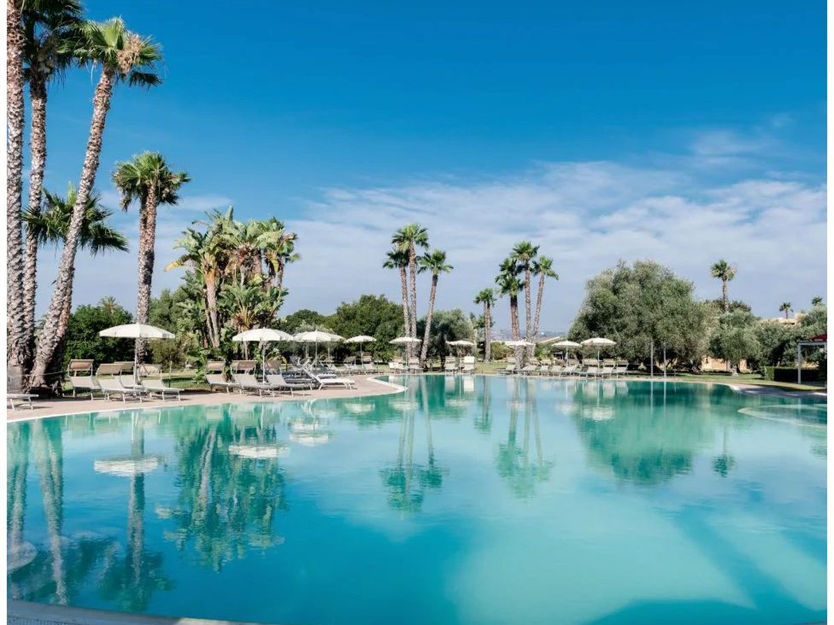 A large curved outdoor pool of clear turquoise water, fringed by tall palms and olive trees, with sun loungers and white parasols along the far edge under a deep blue sky.