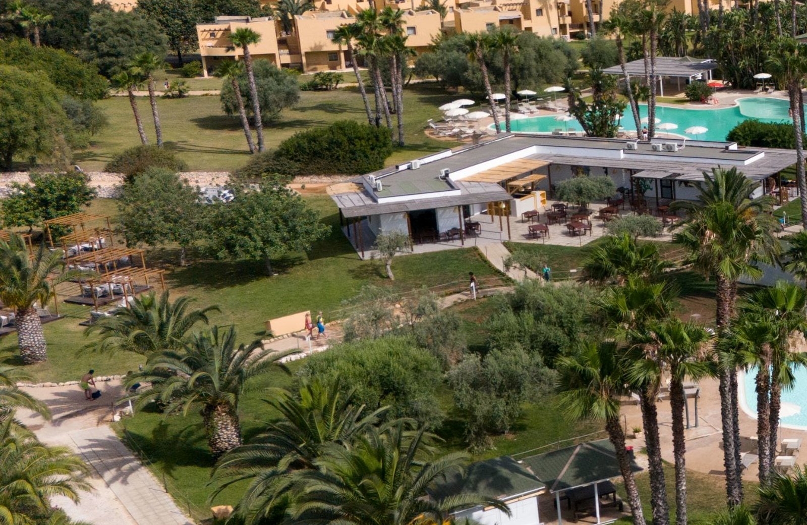 An aerial view over VOI Arenella Resort's parkland, with two-storey ochre guest buildings and palms around a low dining building and its open terrace, and the pools beyond.