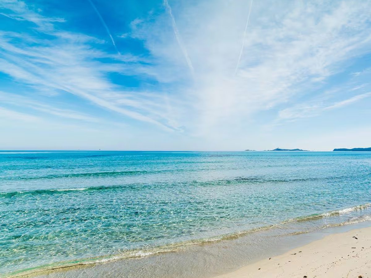 Clear shallow turquoise water washing onto pale sand under a blue sky with wispy cloud, and a small island on the horizon.