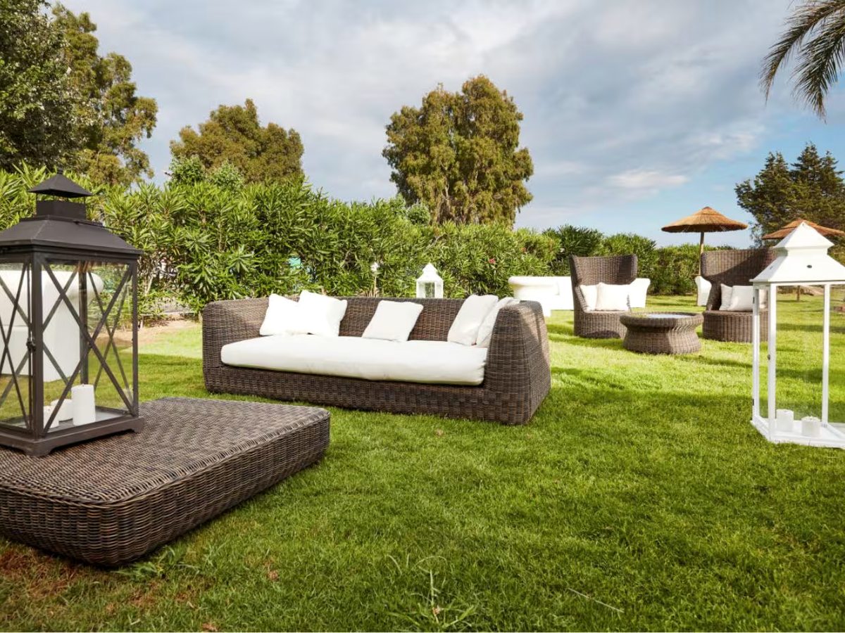 Wicker sofas with white cushions and tall glass lanterns set out on a lawn, with hedges, mature trees and thatched parasols behind.