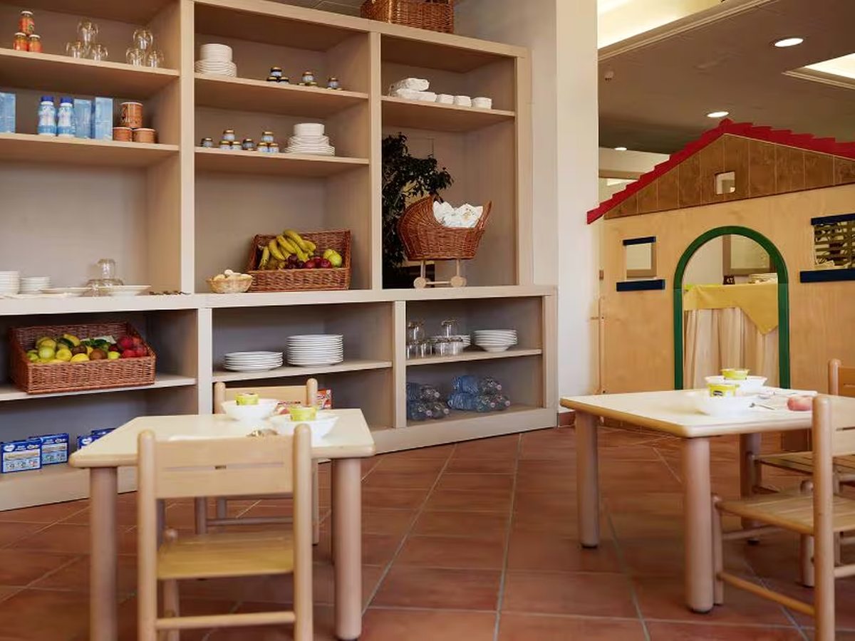 A children's dining room with child-height wooden tables and chairs, shelves of fruit, bottled water and crockery, and a wooden playhouse in the corner.