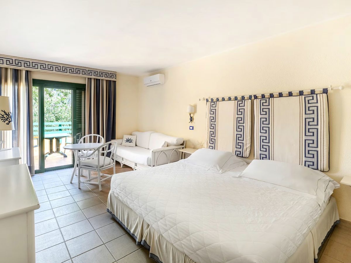 A cream room with two blue and white patterned headboards, a white sofa, a small table and chairs and doors onto a green-railed terrace.