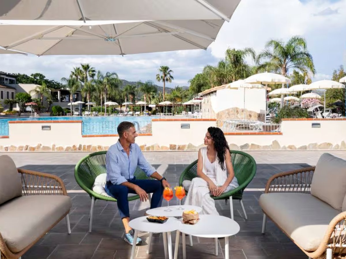 A shaded lounge terrace with wicker sofas and green chairs where a couple sit with drinks, and a large rectangular pool and palm trees behind.