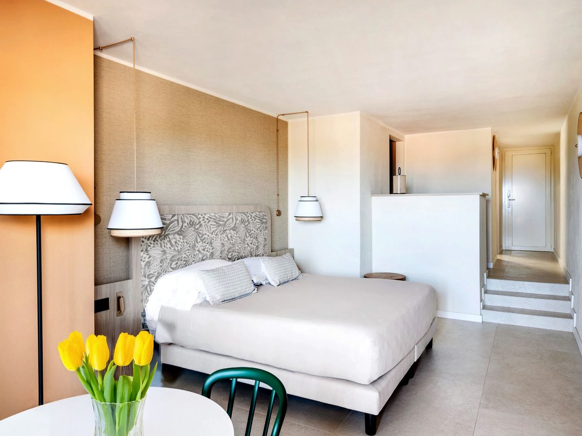 A renovated room in beige and terracotta with a patterned headboard, pendant bedside lights, a floor lamp, a small table of tulips and steps up to the entrance hall.